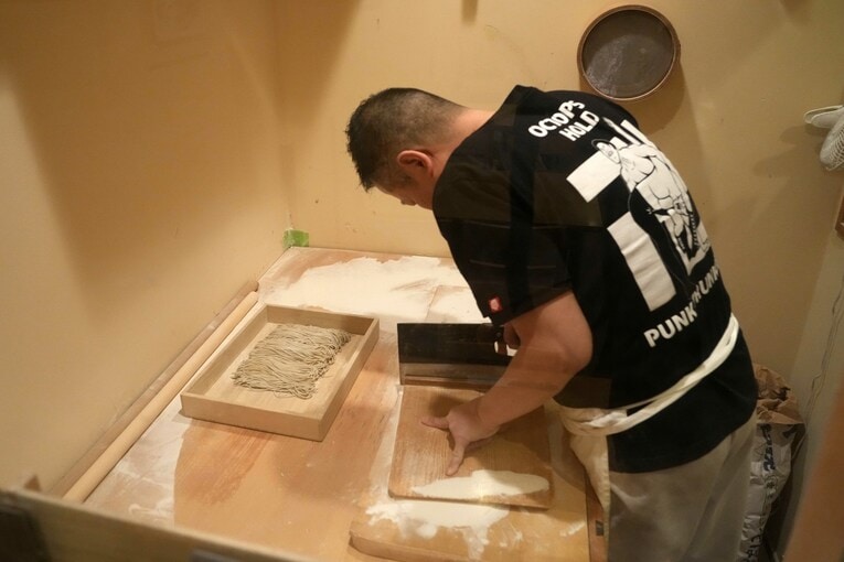 卍固めのTシャツ姿で蕎麦を切る川嶌さん