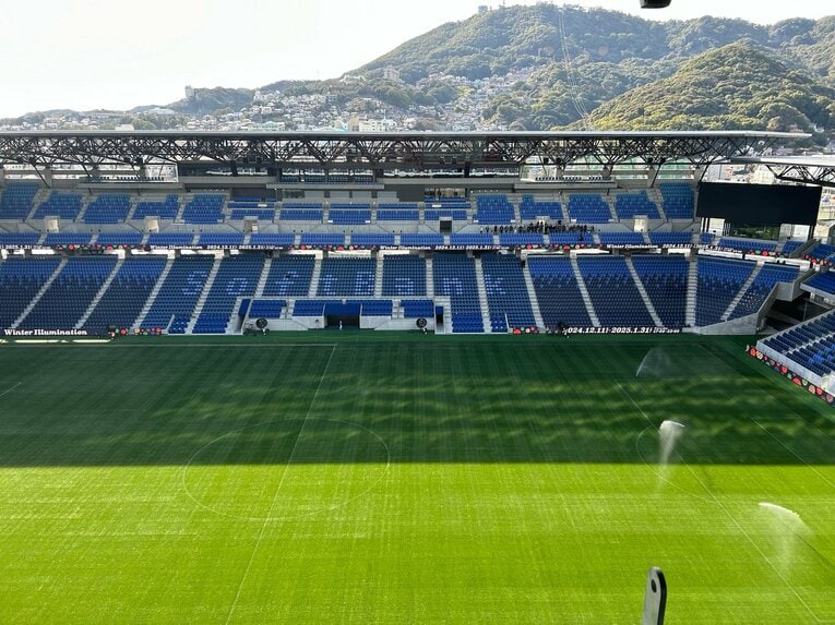 スタジアムのスタンド、緑のピッチが広がり、その先には稲佐山の山頂が見える