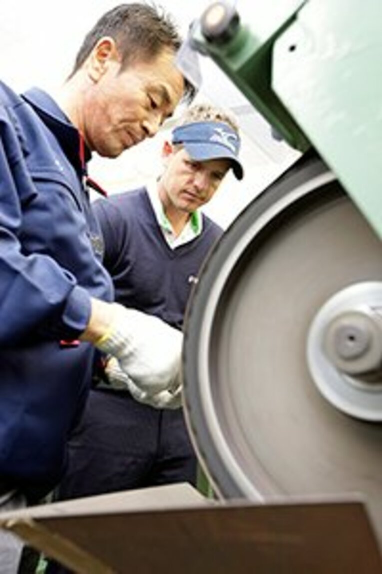 ミズノのアイアンを手作りで仕上げる熟練のクラフトマン。中でもこの道三十余年の伊藤友男さんは国内外のツアープロから絶大な信頼を寄せられている。その伊藤さんの技術を、真剣に見つめるルーク・ドナルド選手。 