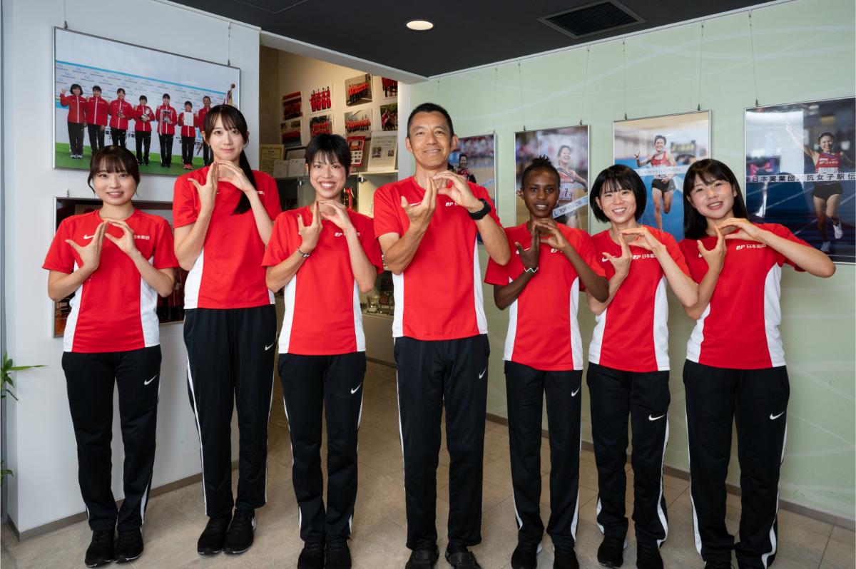 「JP」ポーズをとる、（左から）菅田雅香、牛佳慧、廣中璃梨佳、髙橋昌彦監督、カリバ・カロライン、鈴木亜由子、太田琴菜　©Tadashi Hosoda