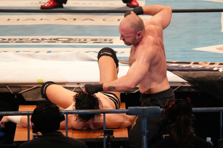 テーブルの上の成田蓮を殴りつけるジョン・モクスリー　©Essei Hara