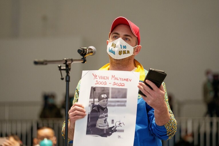 ウクライナ紙キエフ・ポストのリー・リーニー記者。ハリコフで爆撃によって亡くなった19歳のバイアスロン選手の写真を掲げ、パーソンズ会長にロシア選手の“除外”を訴えた　©AFLO