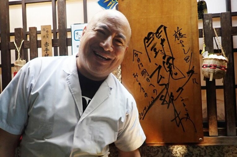 アントニオ猪木が書いた「藤よし」の看板の前で笑顔を見せる「サダさん」こと店主の早川禎行さん ／ photograph by Essei Hara