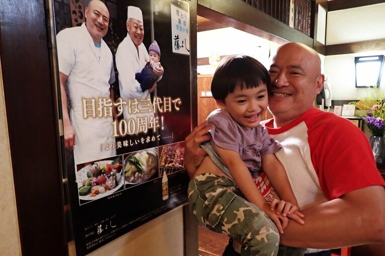 店のポスターの前で長男・寛太郎くんを抱き上げる「サダさん」こと早川禎行さん　©Essei Hara