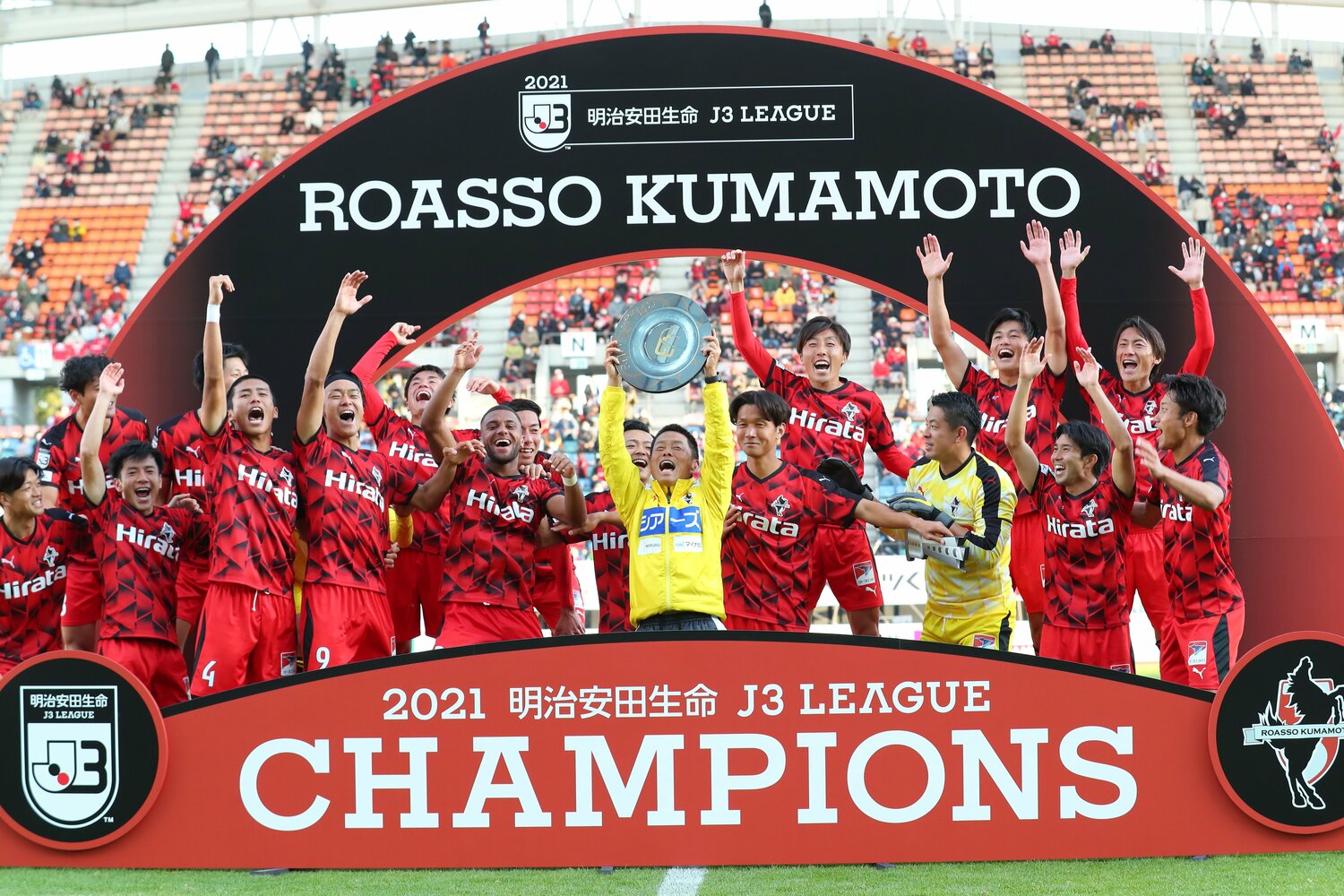 最終節に勝利し、逆転でJ2昇格を決めたロアッソ熊本。選手に囲まれた大木監督は笑顔でシャーレを掲げた　©︎J.LEAGUE